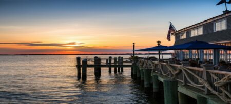 A sunset over calm water, reflecting orange and blue hues, with a wooden pier and restaurant beside it.