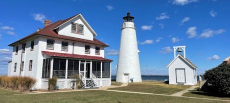 A white lighthouse next to a two-story house with a red roof, and a small bell tower. The sky is blue with clouds.