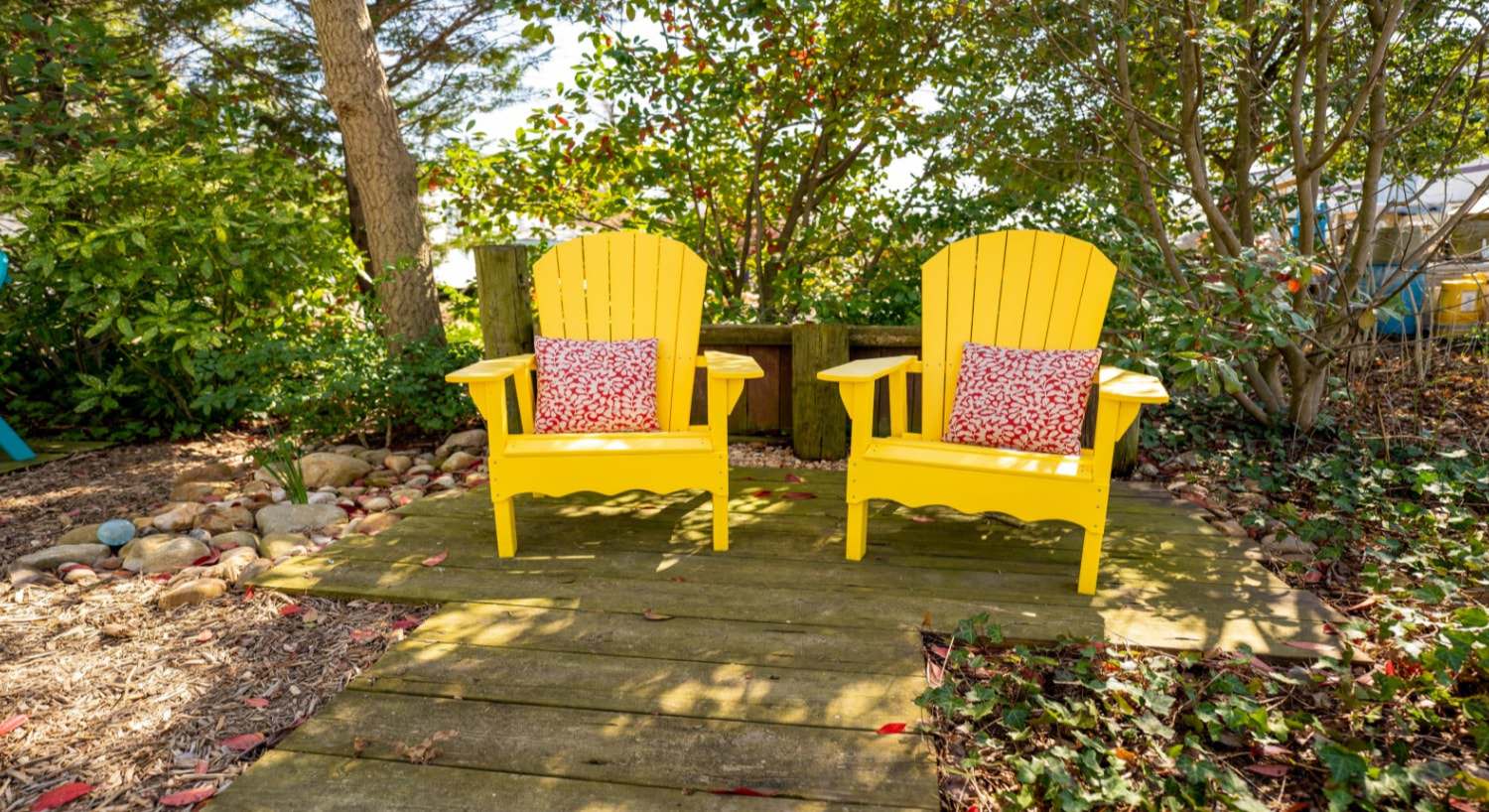 Two bright yellow adirondack chairs each with a red and white floral patterned pillow surrounded by green shrubs and trees