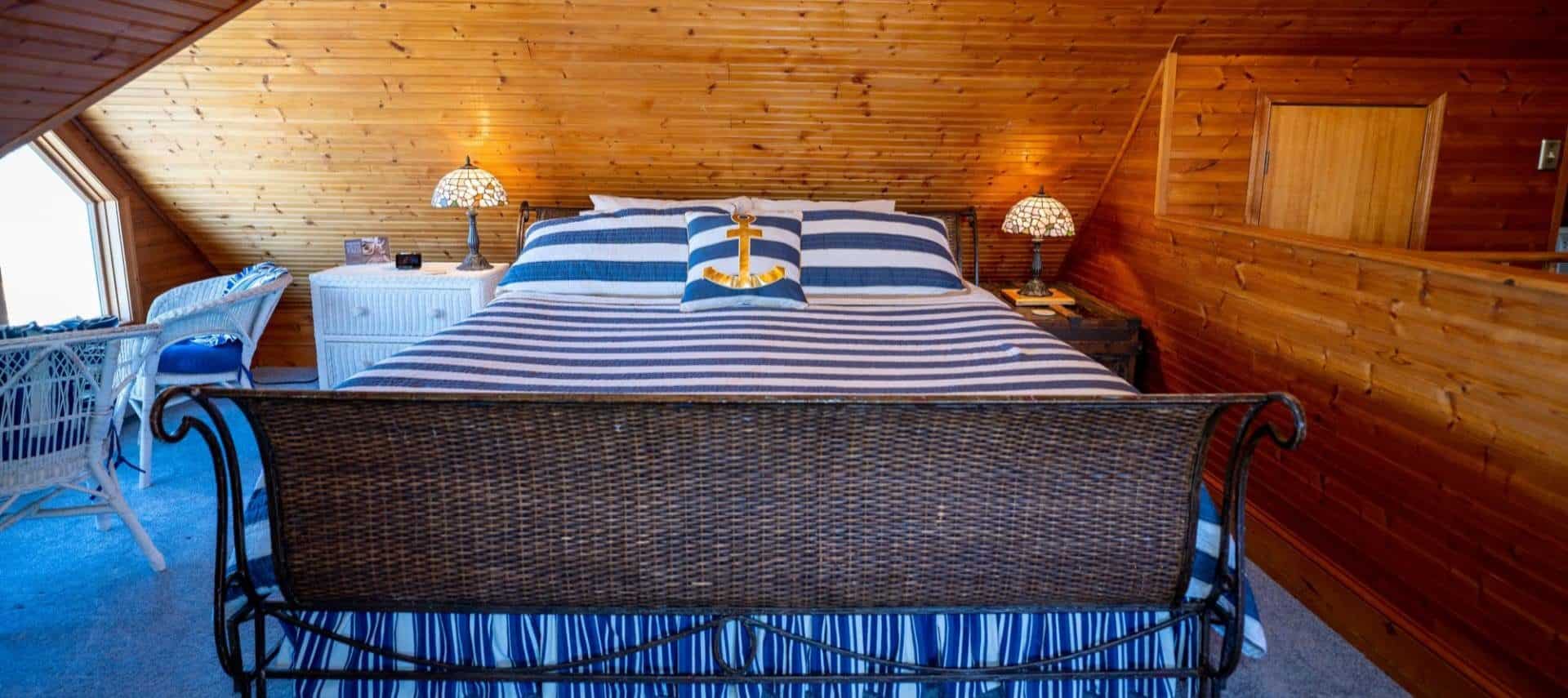 Room with floor to ceiling wood paneling, blue carpeting, dark brown wicker sleigh bed with blue and white striped bedding, and white wicker dresser
