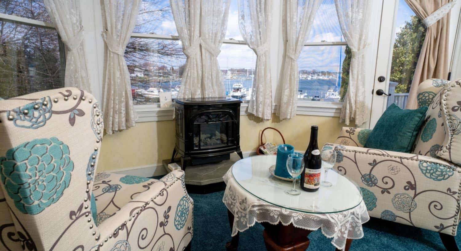 Two upholstered arm chairs with a bold teal and brown flower pattern sitting next to a small table with glass top near a fireplace next to large windows with a view of the marina