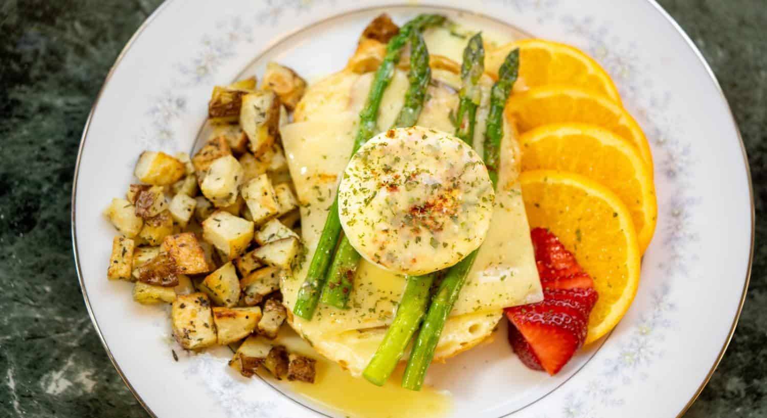 Close up view of eggs benedict dish topped with asparagus with country potatoes on one side and sliced strawberries and oranges on the other side