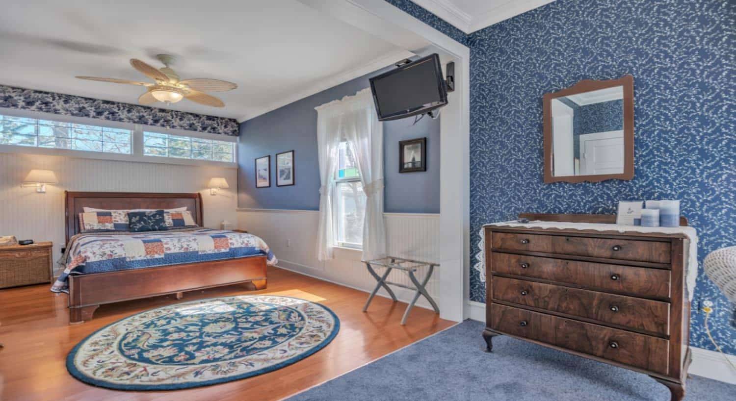 Bedroom with blue-gray walls, white wainscoting, hardwood flooring, dark wooden furniture, ceiling fan, and TV mounted on the wall
